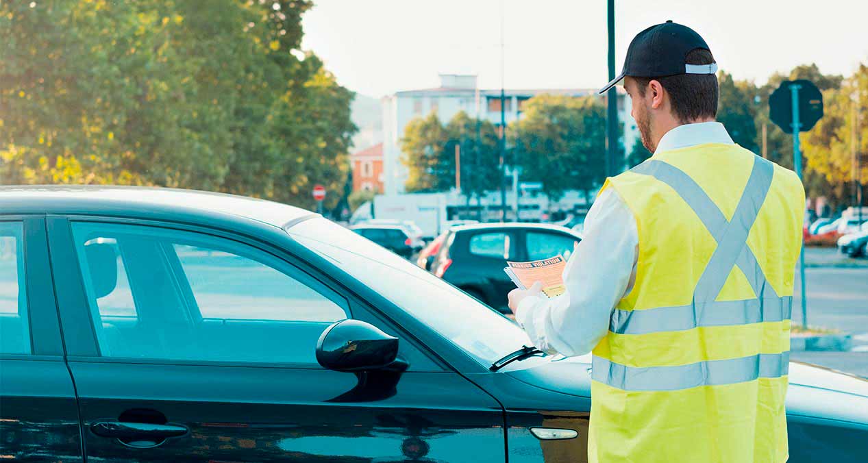 Hombre con chaleco amarillo y gorra coloca un aviso de pago vehicular en un auto 