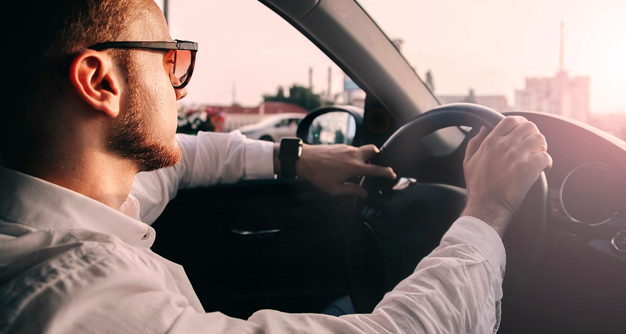 perfil de un hombre de camisa blanca y lentes oscuros manejando un auto al atardecer